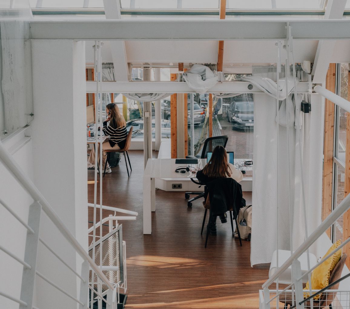 Blick von einer Treppe in ein helles, modernes Büro mit Holzfußboden, Glasfronten und zwei Personen, die an Schreibtischen arbeiten
