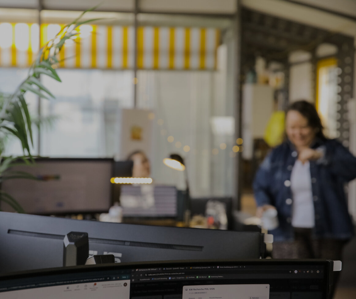 Unscharfe Büroszene mit Monitor im Vordergrund und einer Person mit Tasse im Hintergrund