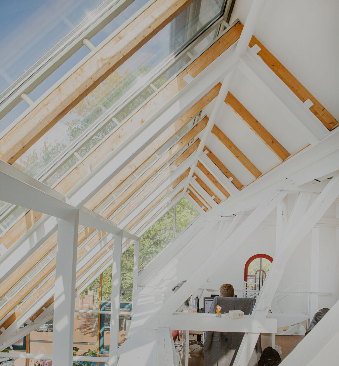 Helles Dachgeschossbüro mit großen Glasdachfenstern, sichtbaren Holz- und Stahlträgern und einer Person am Schreibtisch