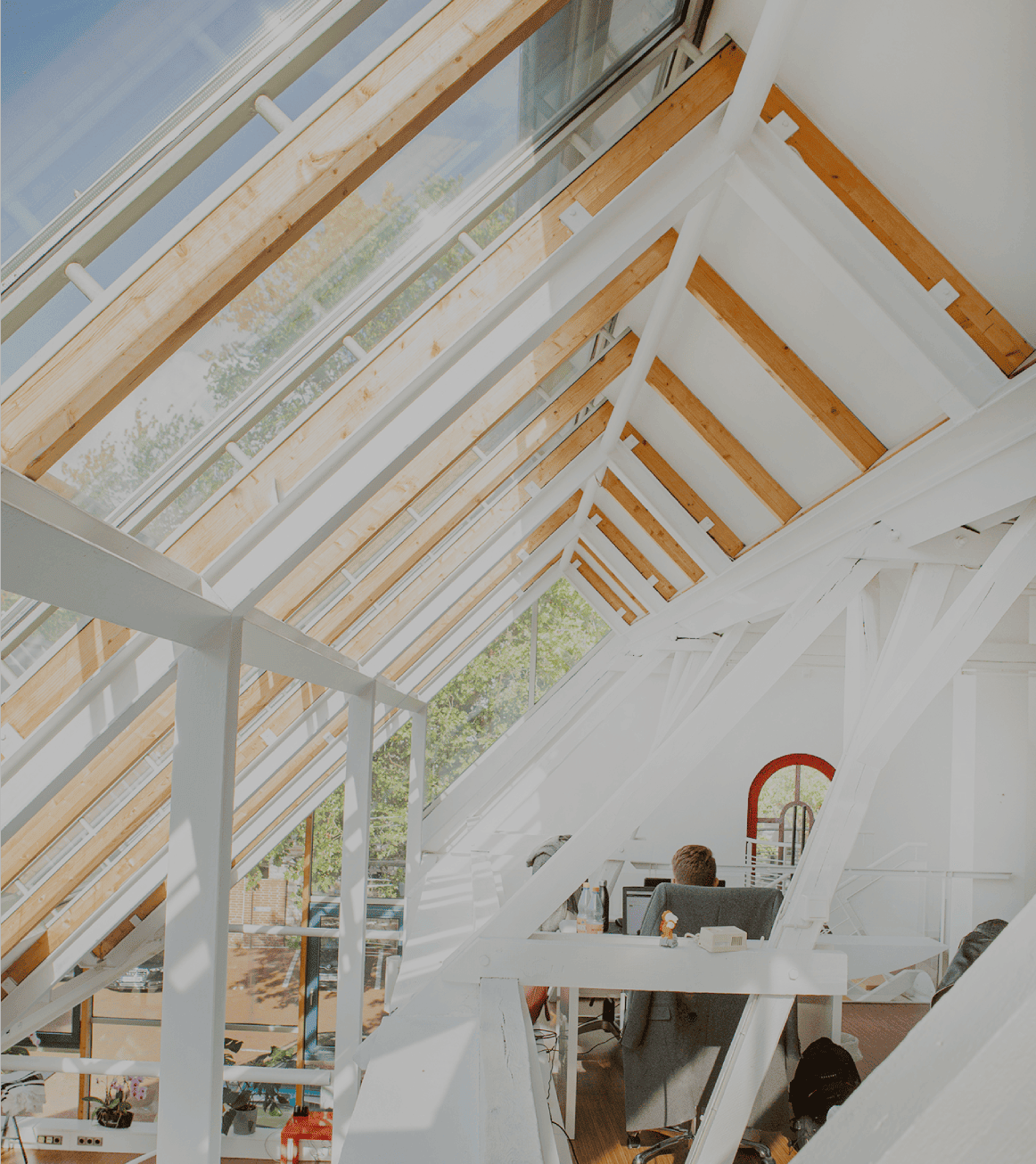 Helles Dachgeschossbüro mit großen Glasdachfenstern und sichtbaren Holz- und Stahlträgern; eine Person sitzt an einem Schreibtisch.
