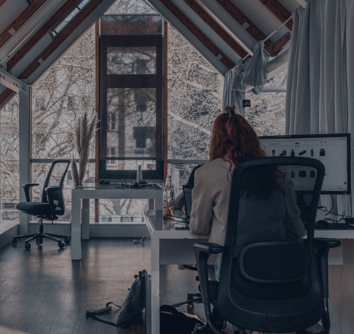 Person am Schreibtisch in einem hellen Büro mit großen Dachfenstern und Monitor