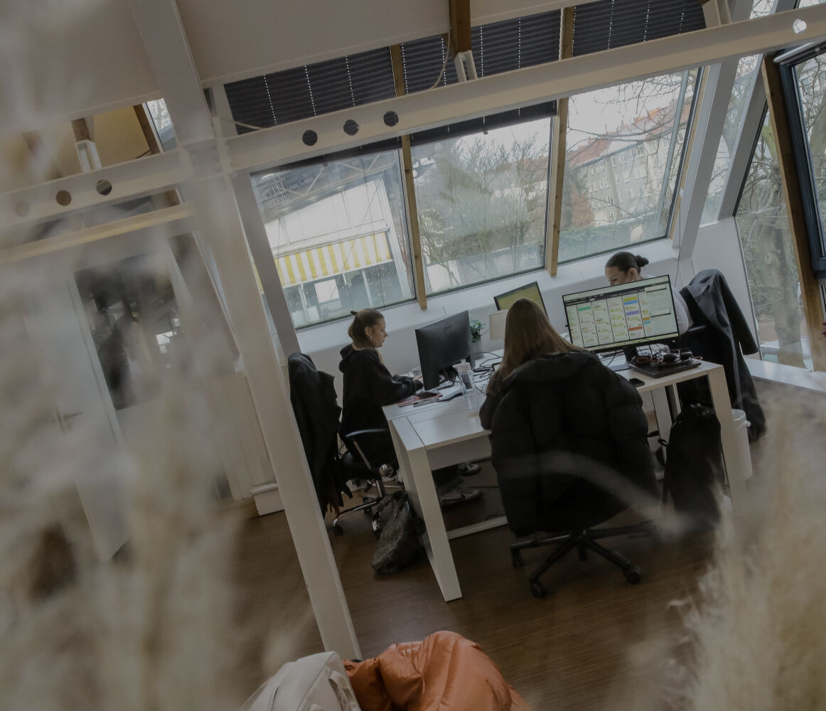 Person arbeitet an einem Schreibtisch im Büro, seitlich durch eine Glaswand fotografiert, mit Fensterfront im Hintergrund.