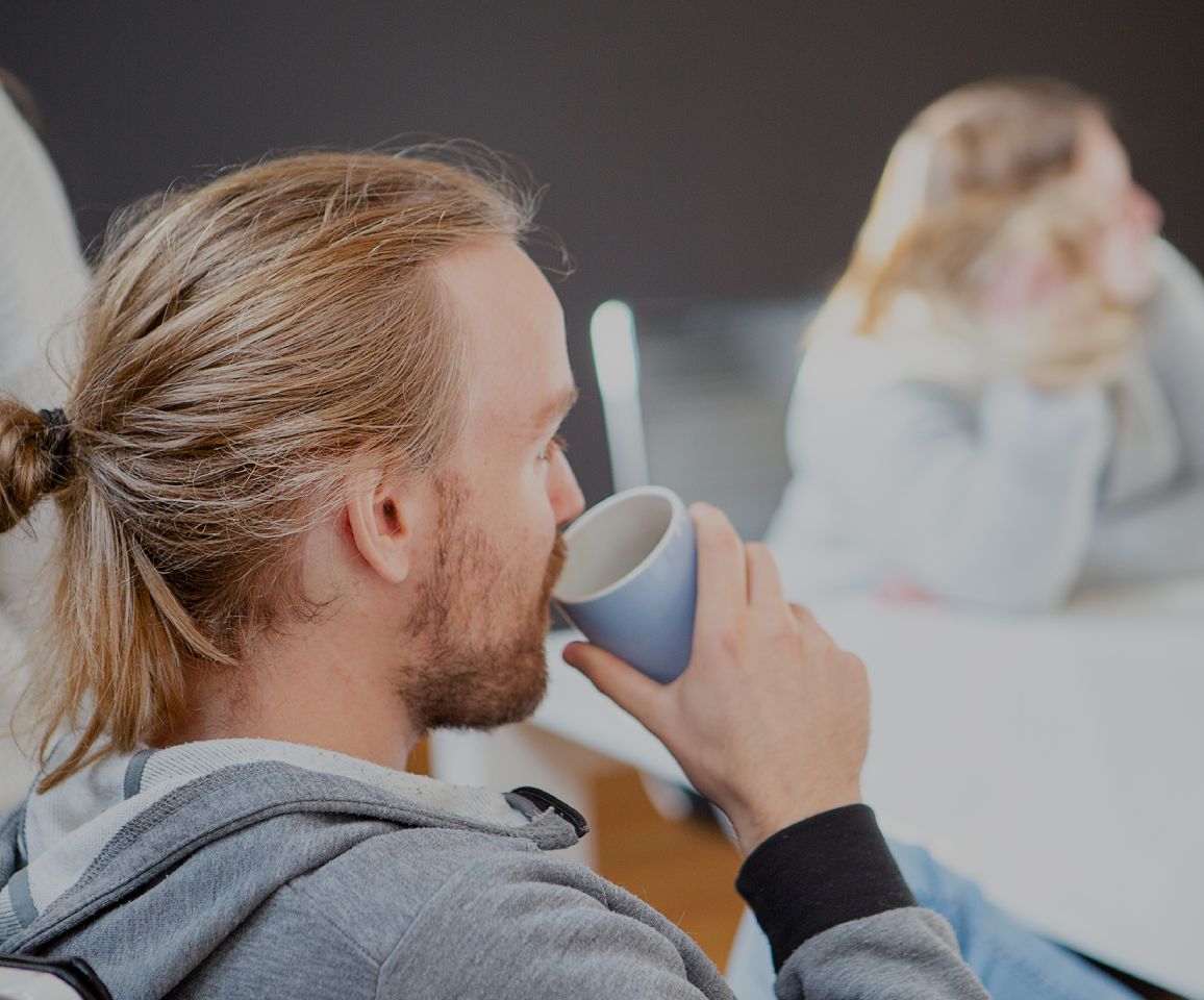 Person mit zusammengebundenem Haar trinkt aus einer Tasse in einem Besprechungsraum; im unscharfen Hintergrund sitzt eine weitere Person am Tisch, helle, entspannte Büroszene