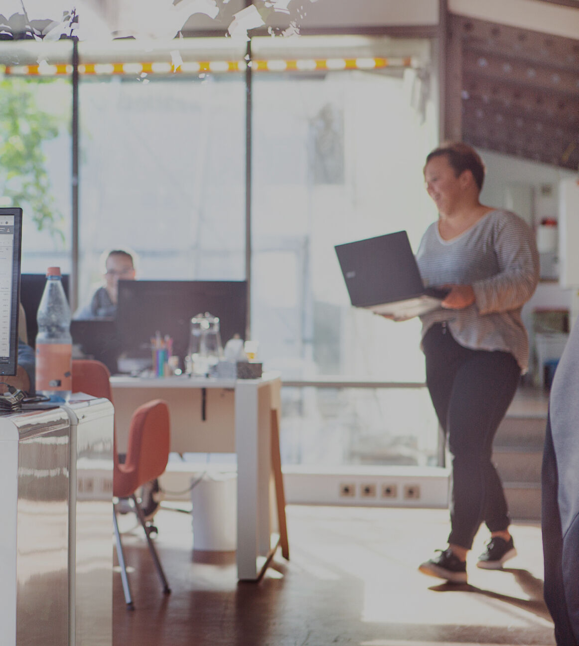 Heller Büroraum mit mehreren Arbeitsplätzen, im Vordergrund eine Person am Schreibtisch vor einem Monitor mit Tabellenansicht