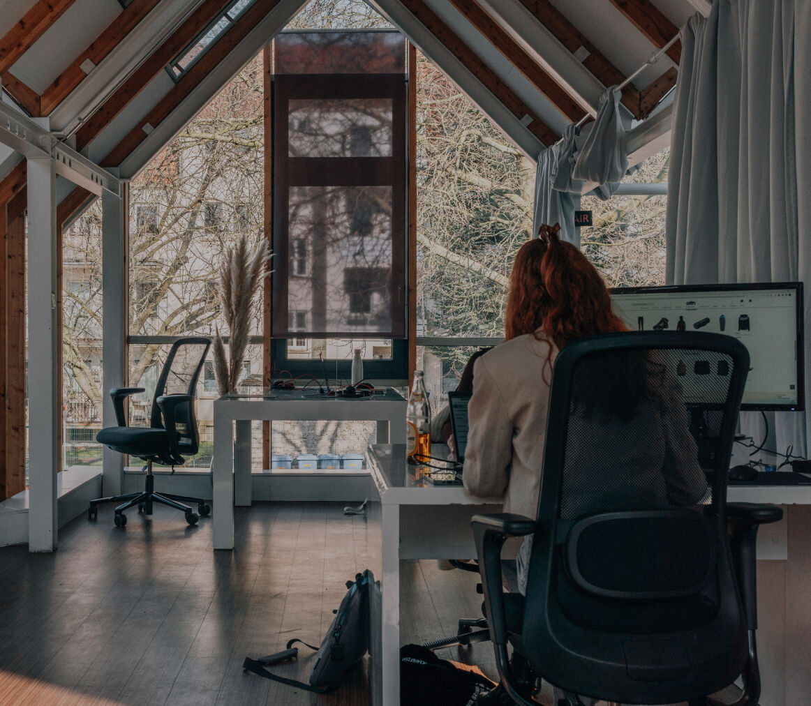 Zwei Personen arbeiten an separaten Schreibtischen in einem hellen Büro mit großen Fenstern und sichtbaren Holzbalken